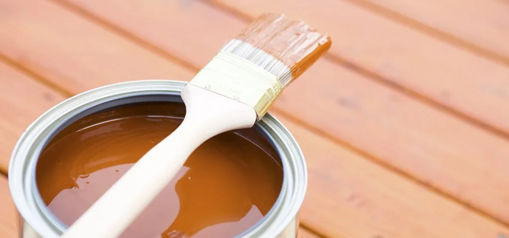 A close-up photo of a paint brush resting on top of a can of brown paint. The can and brush sit on a wooden deck surface.